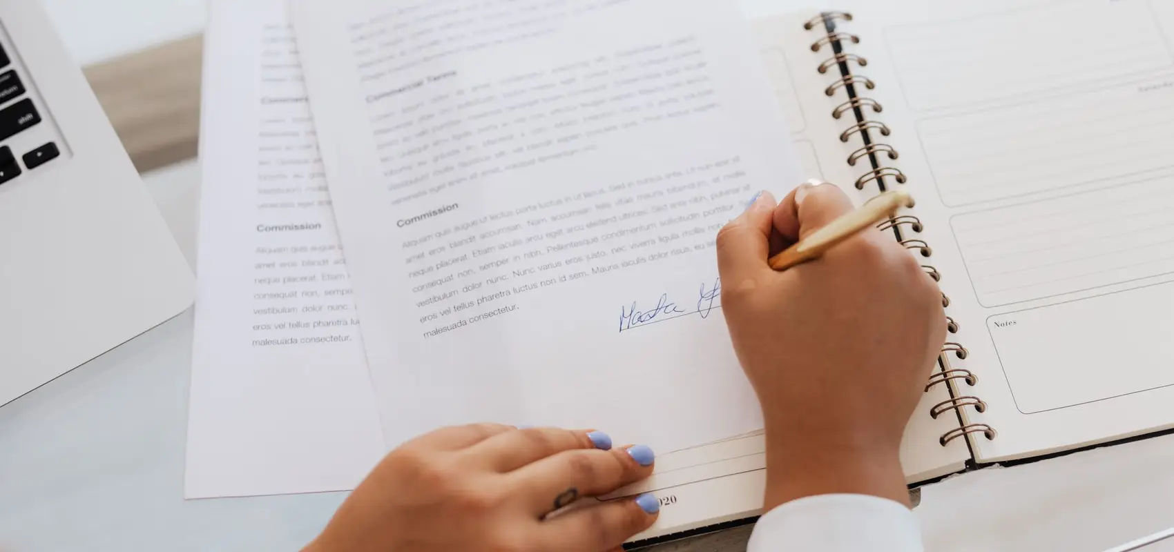 Main signant un contrat imprimé posé sur un carnet, à côté d’un ordinateur portable